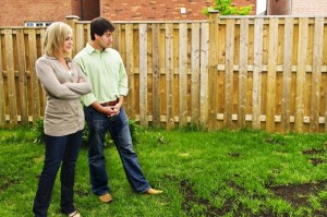 A blond woman in cream-colored jacket and jeans stands to the left of a man in short-sleeved light green button-down shirt and jeans, staring at a bare patch in the grass in their backyard, with concerned looks on their faces. Wooden fence in background.
