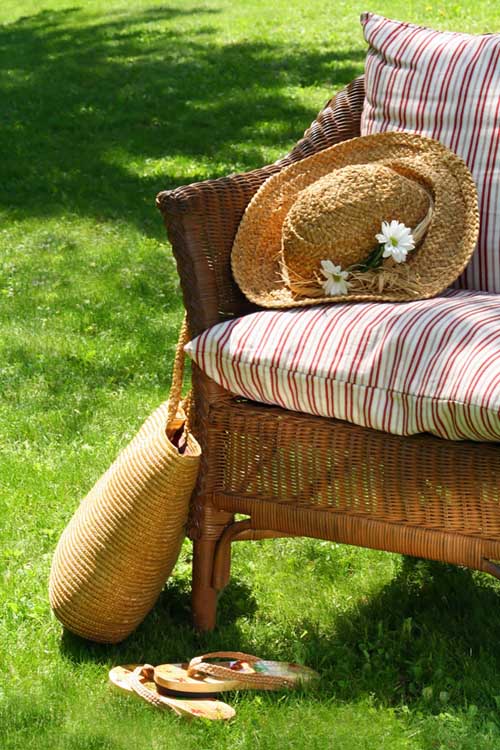 A close up picture of a wicker armchair with striped cushions set on a lawn on a sunny day, with a straw hat and tote bag to the left.