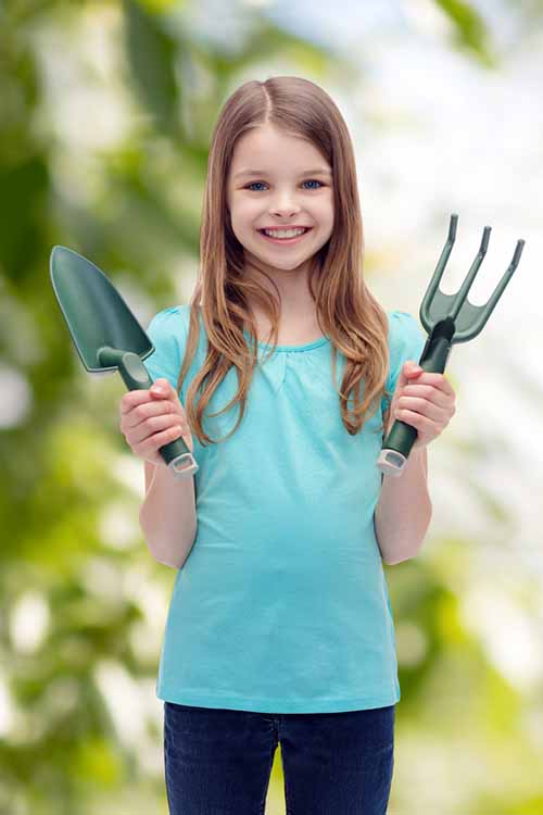 A close up vertical picture of a young girl holding gardening tools, on a soft focus background.