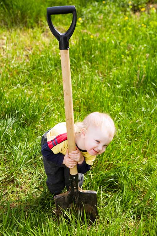 A close up of a toddler smiling and playing on a lawn with a large garden shovel, in bright sunshine.
