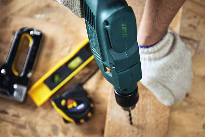 A close up of a green electric drill making a hole in a piece of timber held down by a gloved hand. In the background is a spirit level and further household tools in soft focus.