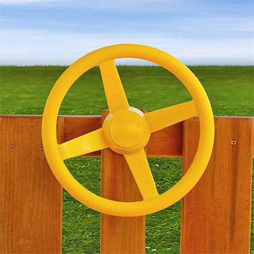 A close up of the Gorilla Playsets Steering Wheel, in yellow, attached to a wooden fence, with lawn in the background.