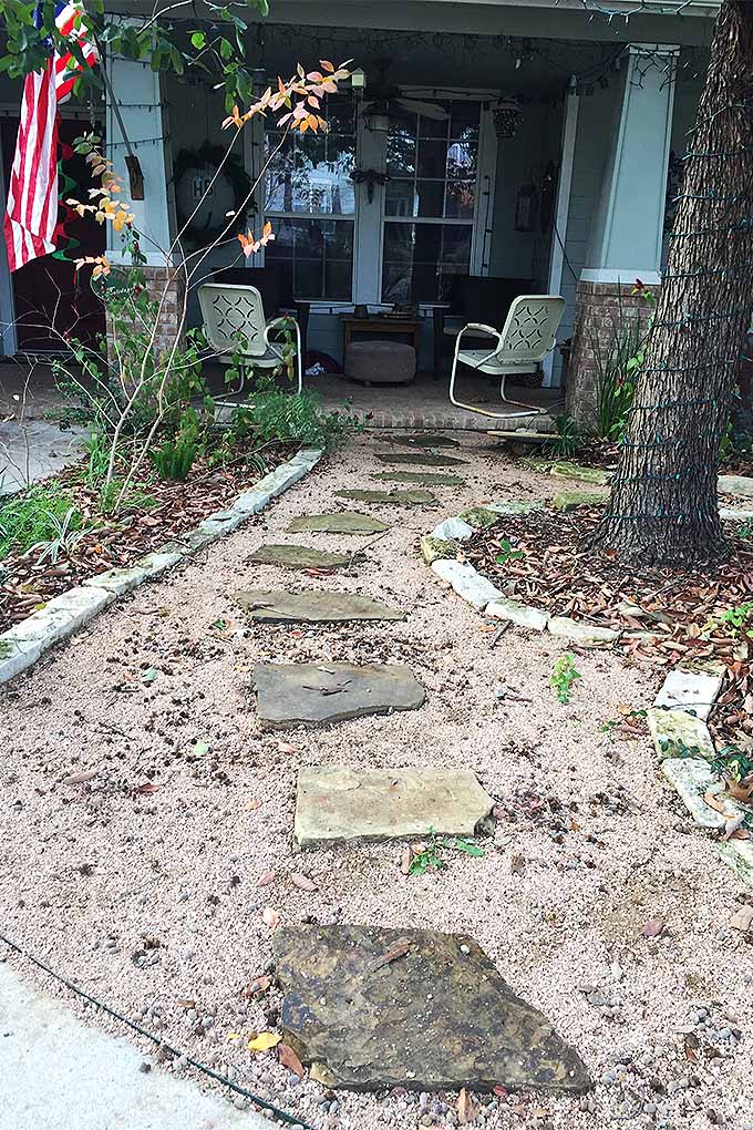 A vertical image of a garden walkway made with stepping stones embedded in crushed granite and edged with bricks, leading to a porch at the front of a suburban home