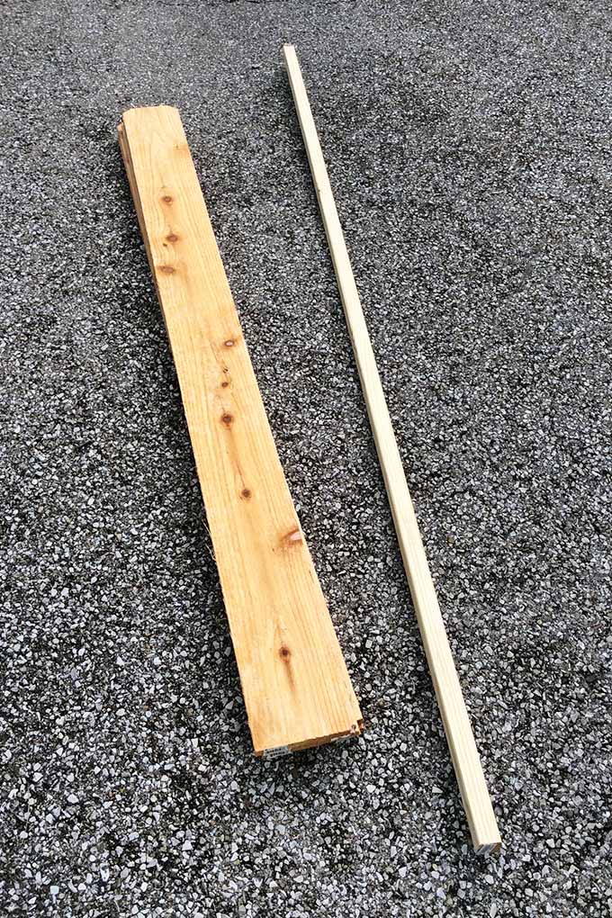 Vertical top-down view of a piece of lumber beside a measuring stick or a narrow piece of wood, on a blacktop driveway.