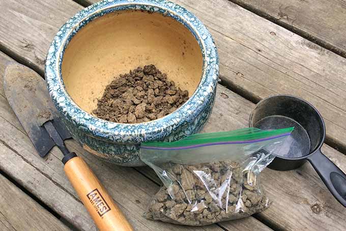 A small handheld spade sits on a rough wooden table next to a ceramic pot and a bag both containing samples of dirt. The soil in them is dry and clumped together. To the right of them all is a small, black measuring cup.