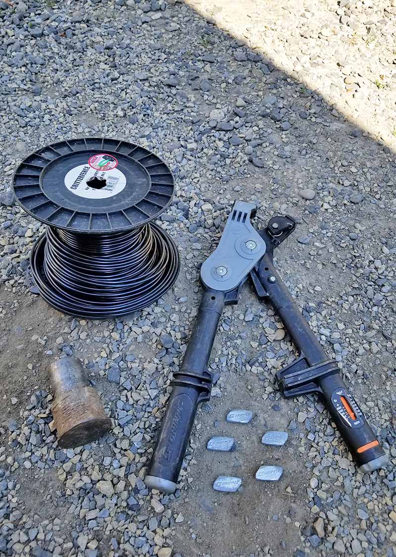 A ratcheting tool, spool of black tension wire, metal Gripples, and other implements used to install deer fencing, on gray asphalt in partial shadow.