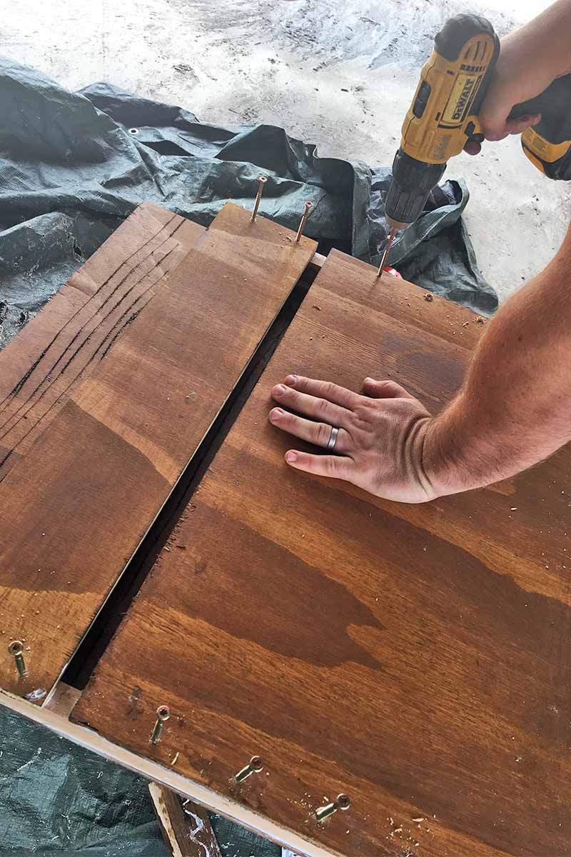 Closeup of a man drilling screws into a brown stained wooden box with one hand while holding it steady with the other, on a black tarp on top of a cement floor.