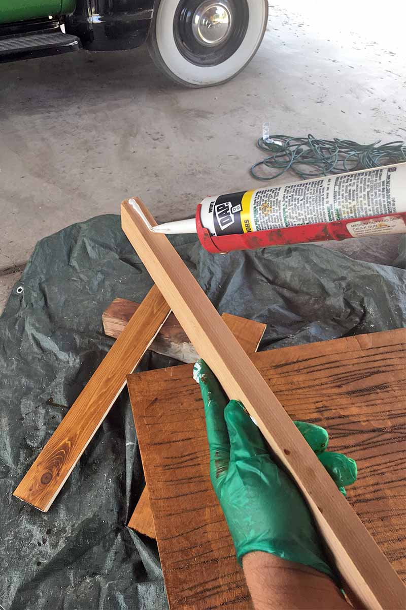 Two hands covered with green gloves hold a thin strip of wood in the left and run a bead of caulk down the center with the right, with brown stained wood and a black tarp on a cement floor in the background, in front of a green vehicle with white-wall tires.