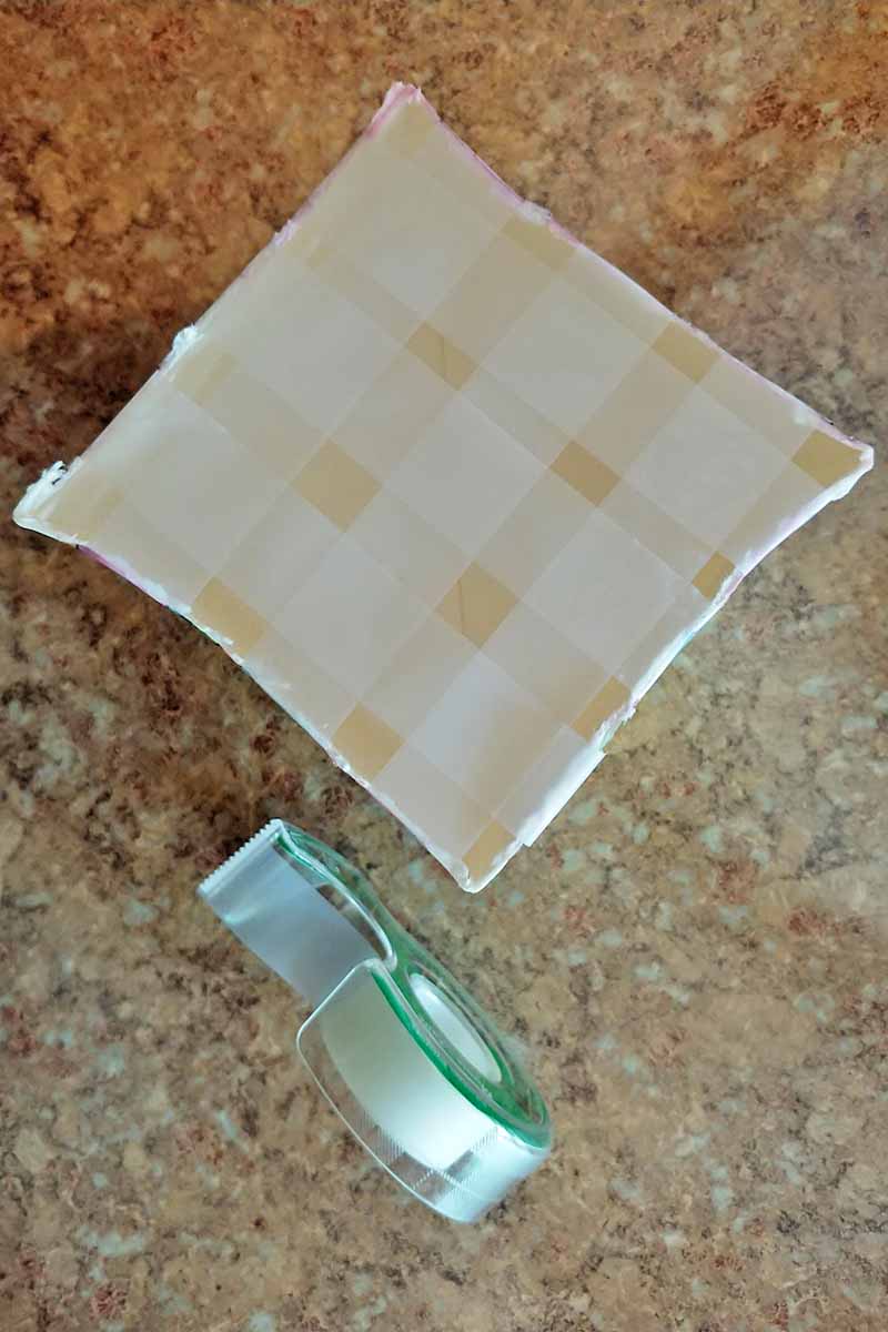 Overhead vertical shot of a roll of Scotch tape and a half-gallon milk carton that has a grid of tape on the open top, on a light brown surface.
