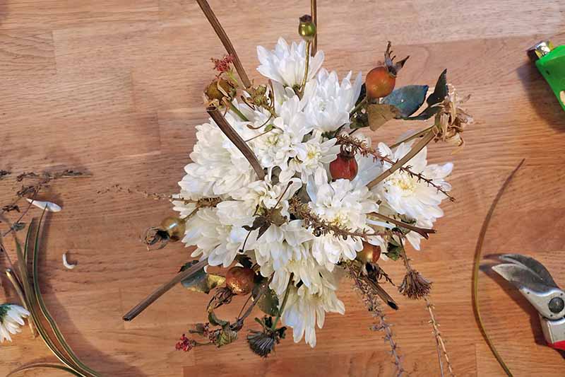 Overhead shot of a flower arrangement with white mums and various types of greenery from the garden, with more to the left on a wood surface, and garden pruners to the right.