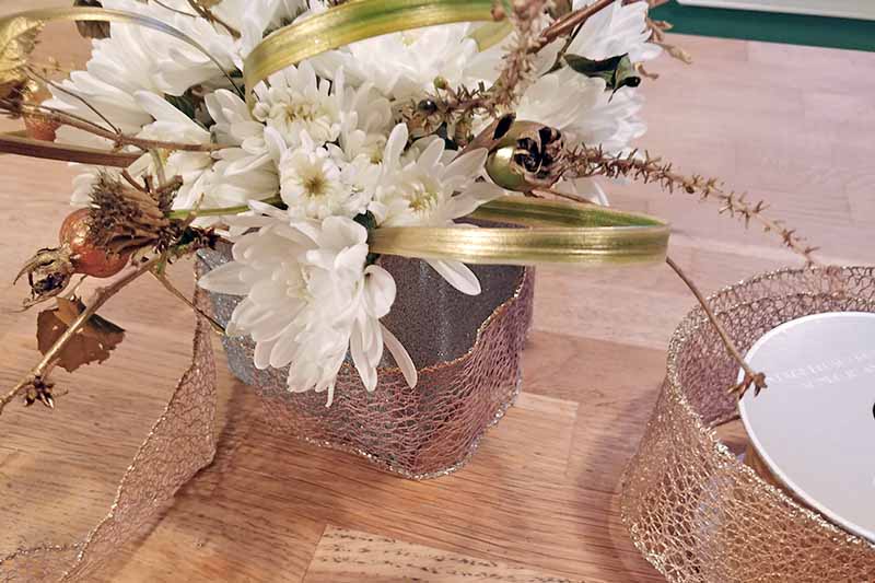 Closeup closely cropped image of a flower arrangement with white mums, greenery from the garden, and loops of ornamental grass, in a square silver spray painted container wrapped loosely in wired decorative ribbon with more on a spool to the right, on a brown wood surface.