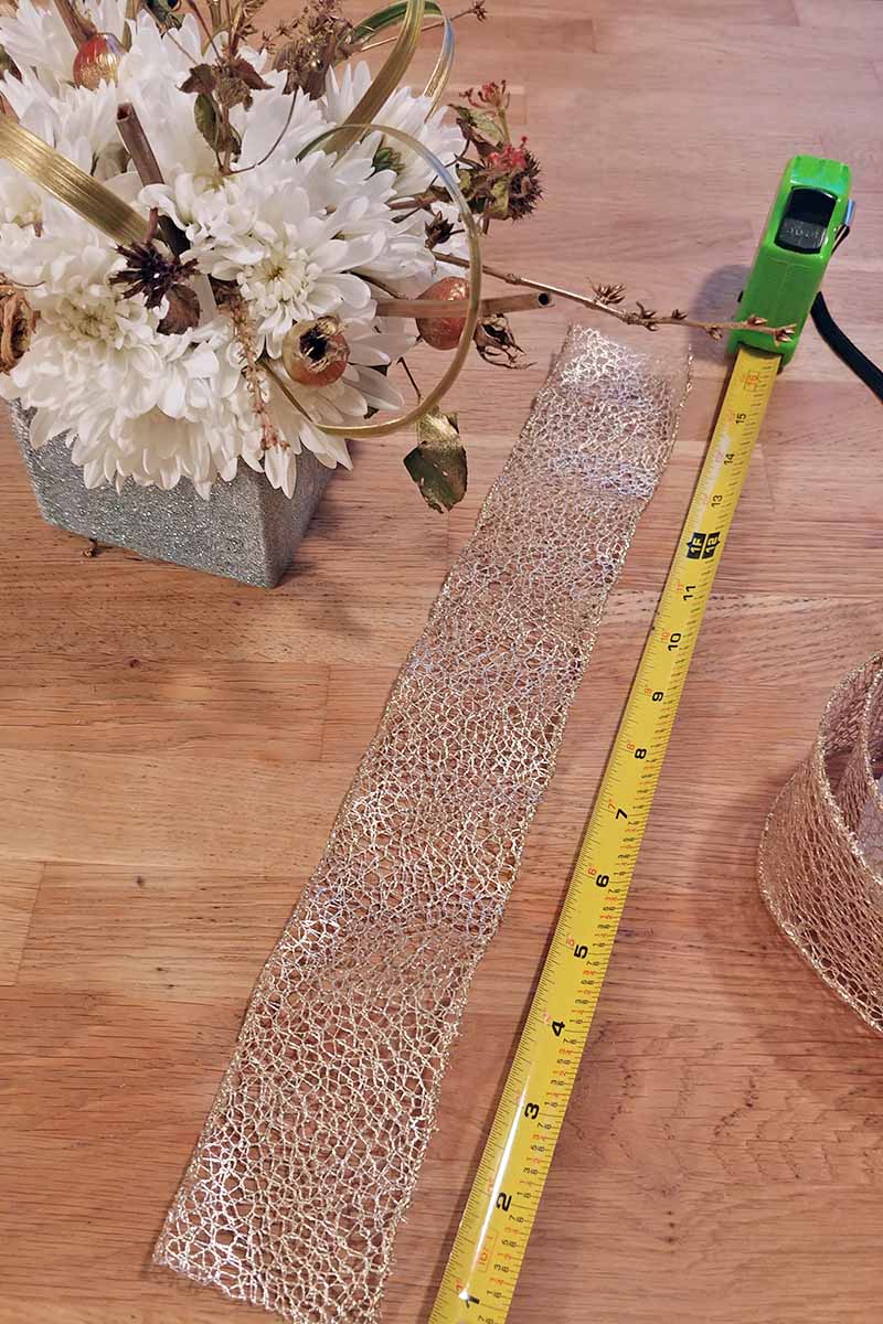 A yellow tape measure with a green plastic case is being used to measure decorative wired ribbon on a wood surface, with more ribbon to the right and a flower arrangement to the left.