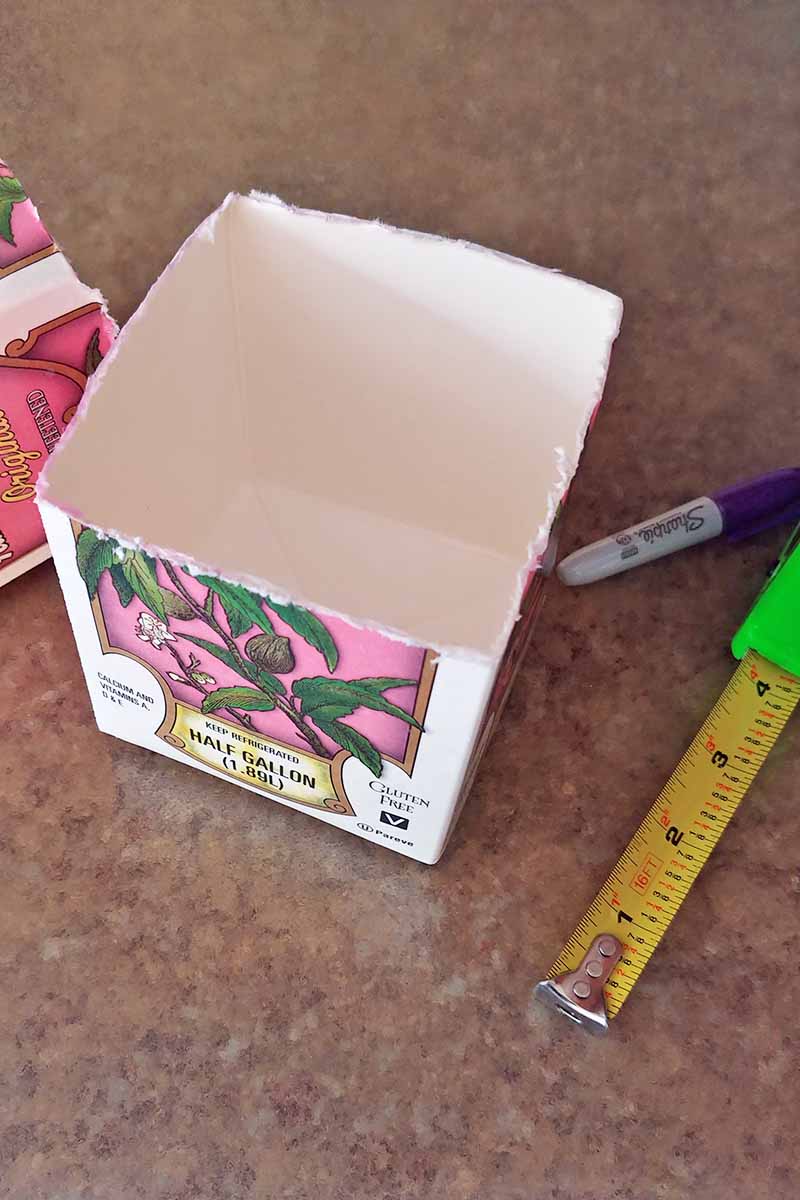 Half of a cardboard almond milk carton with a rough edge, with the removed top to the left, and a purple Sharpie market and yellow and green tape measure to the right, on a beige countertop.
