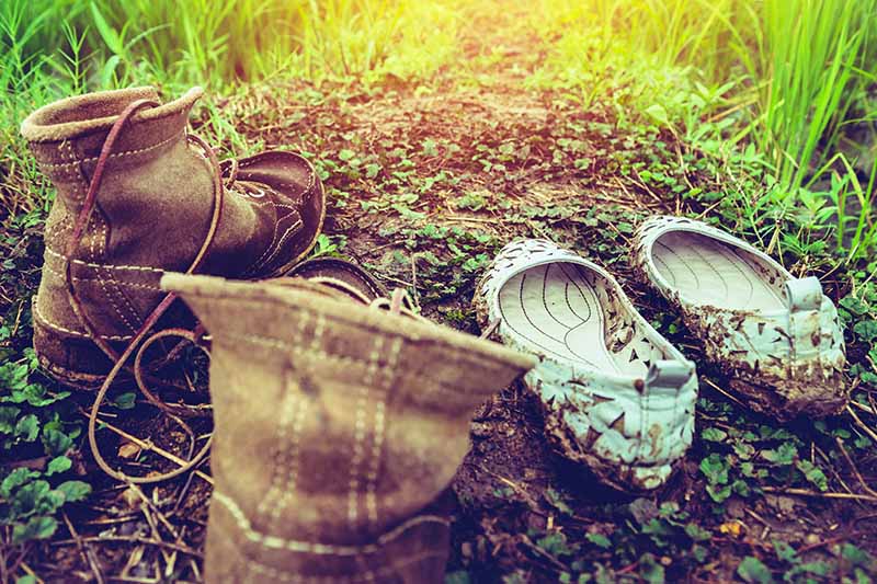 Horizontal image of worn brown boots and mud-covered ballet flats, in brown soil with green glass, and bright filtered sunlight.