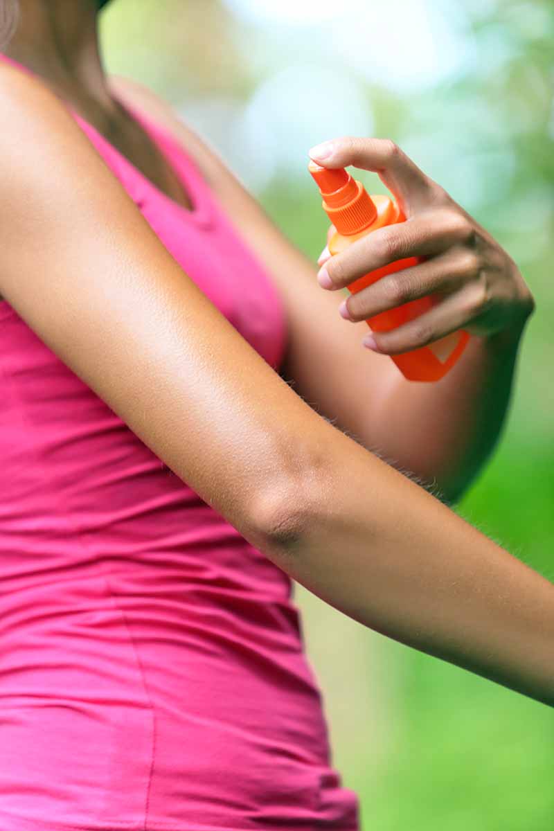 Closely cropped image of a woman's torso, clothed in a pink tank top with bare arms, spraying a bottle of orange insect repellent onto her skin.