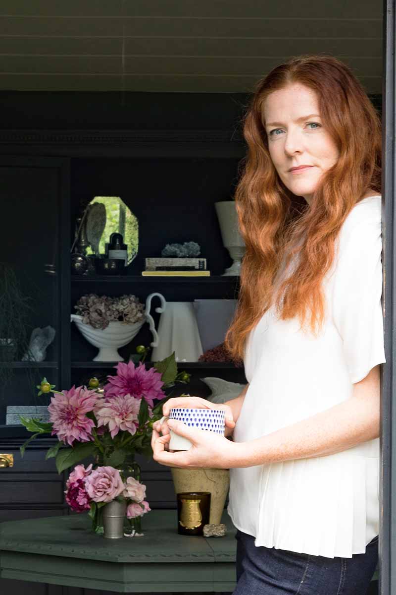 Vertical image of Clare Nolan in a white tee-shirt, with plants and containers on a shelf in the background.