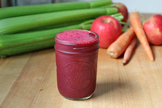 Close up of a jar with fresh beet juice, on a wooden surface. In the background are some celery stalks, apples, and carrots in soft focus.
