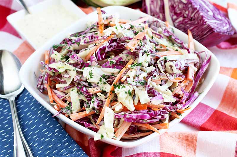 A white bowl with coleslaw made from white and red cabbage, carrots, and herbs. In the background is half a red cabbage. To the left of the frame is a silver spoon on a blue striped cloth, the background is a colorful red, orange, and white tablecloth.