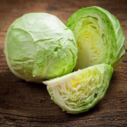 A close up of a cabbage of the 'Late Flat Dutch' variety, a whole one, and one cut in half, with a quarter in the foreground. The background is a wooden surface.
