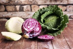 A wooden surface with three different Brassica oleracea vegetables on it. From the left, a white one, chopped in half, in the center a red variety with a slice out of it, and to the right a savoy cabbage, whole.