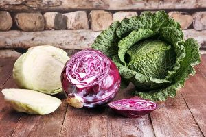 A wooden surface with three different Brassica oleracea vegetables on it. From the left, a white one, chopped in half, in the center a red variety with a slice out of it, and to the right a savoy cabbage, whole. The background is wood.