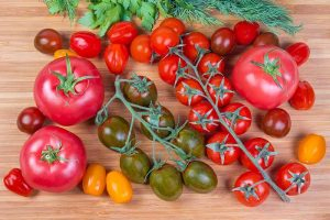 A selection of different heirloom tomatoes shown on a wooden background. A variety of large and small red fruits, as well as yellow and dark green cultivars. Some of the smaller fruits are still attached to the vine, and to the top of the frame are some herbs, just visible.