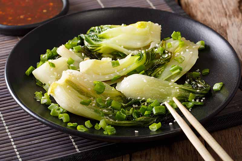 A close up of a dark bowl containing a stir fried dish of bok choy with two chopsticks to the right of the frame. The background is a dark bamboo table mat on a wooden surface.