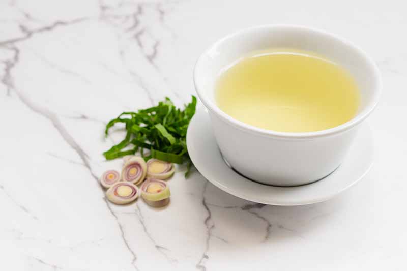 A close up of a white porcelain cup and saucer with a yellow colored tea. To the left of the cup is slices of lemongrass leaves and stalks, on a light marble surface.