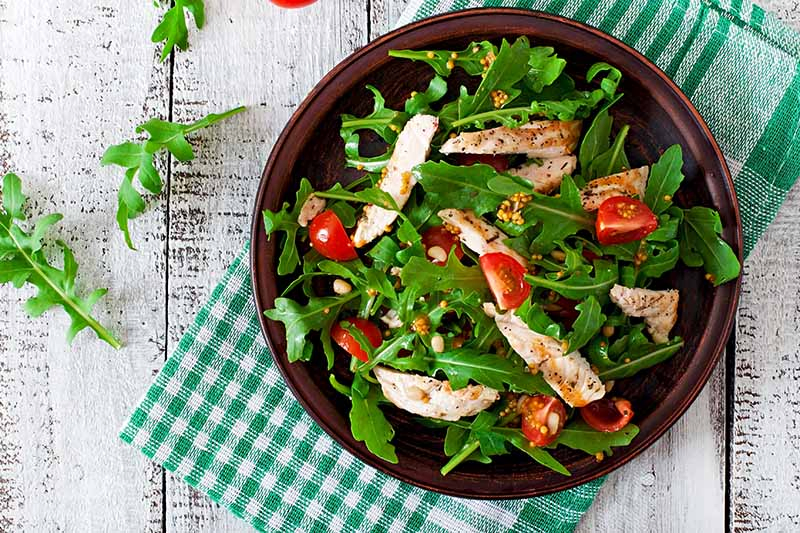 A top down close up picture of a salad of arugula, fresh cherry tomatoes and chicken, set on a green checked cloth on a wooden surface.