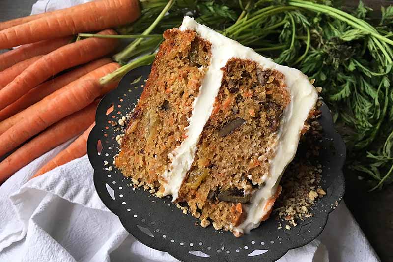 A close up of a dark plate with a slice of carrot cake on it with rich white frosting, set on a white cloth with the vegetables in the background.
