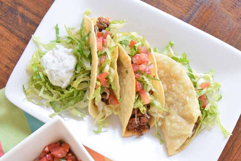 A top down close up of a white rectangular plate with beef tacos and iceberg lettuce set on a brown wooden surface.