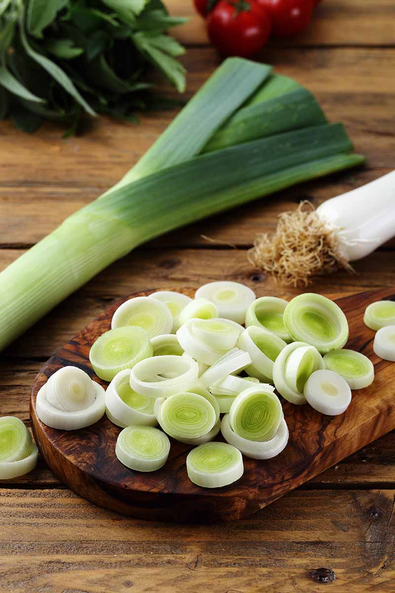 A vertical picture of leeks on a wooden chopping board with some of the stems chopped, and the tops visible in the background, set on a wooden surface.