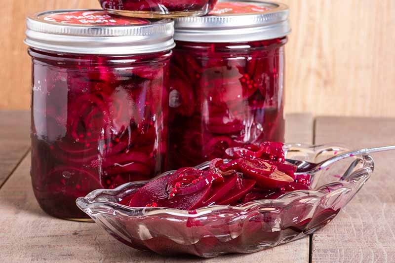 A close up of two jars containing pickled beets with a small glass serving bowl in front of them, set on a wooden surface with a wooden wall in the background.