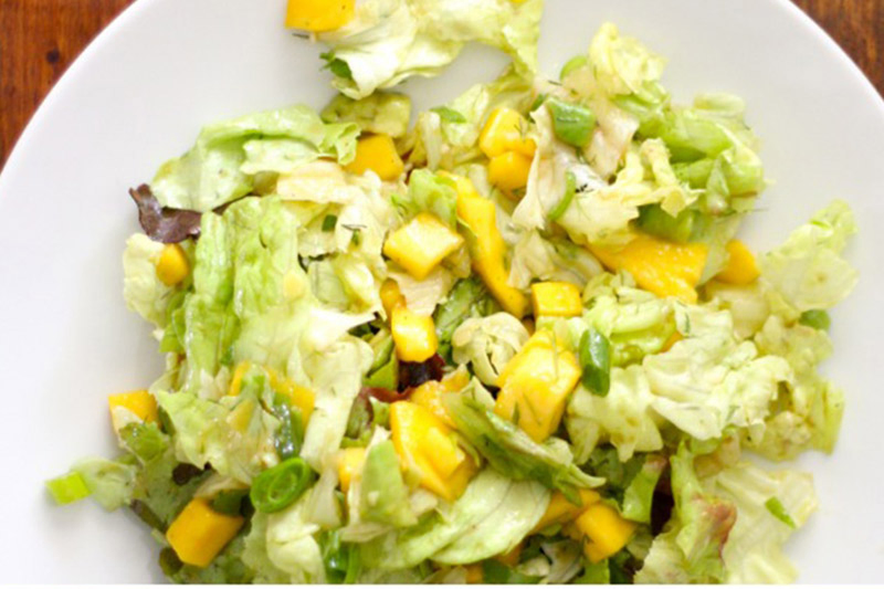 A top down close up picture of a white plate with a mango, avocado, and butterhead salad with tangy dressing set on a wooden surface.
