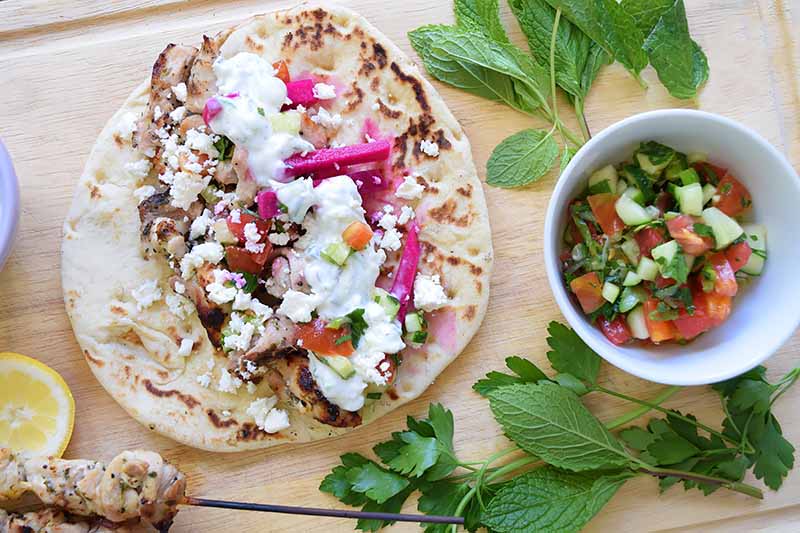 A top down close up picture of a chicken gyros dish, a flatbread with roast chicken, topped with feta cheese and other condiments. To the right is a small white bowl with a cucumber and tomato salad, and herb leaves scattered around. The background is a wooden surface.