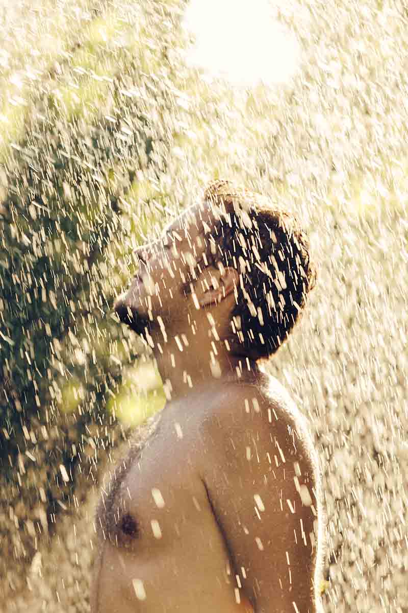 A vertical picture of a young man standing outside with his chin raised and his eyes closed in a shower of rain in bright sunshine.