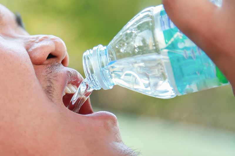 A close up of a plastic bottle pouring water into a man's mouth on a green soft focus background.