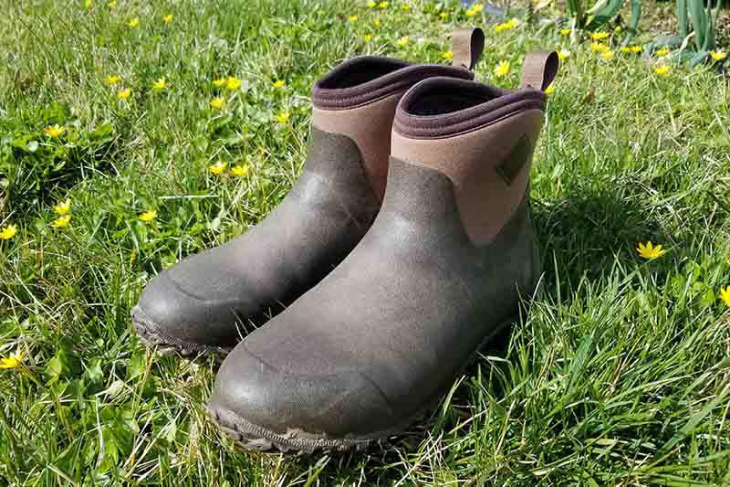 A close up of a pair of the Original Muck Boot Company's Muckster II Ankles in brown, set on a green lawn with small yellow flowers and pictured in bright sunshine.