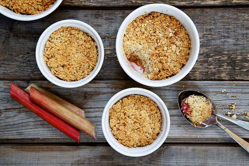 A close up top down picture of four white ceramic bowls containing a breakfast crumble made from freshly harvested rhubarb, to the right of the frame is a spoon and to the left are some fresh red stalks. The background is a rustic wooden surface.