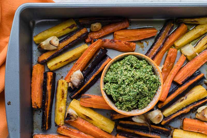 A close up top down picture of a dark gray roasting pan filled with root vegetables and a small bowl of freshly prepared pesto.