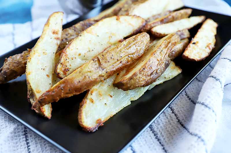 A close up of roasted potato wedges set on a rectangular black plate on a white cloth with black stripes.