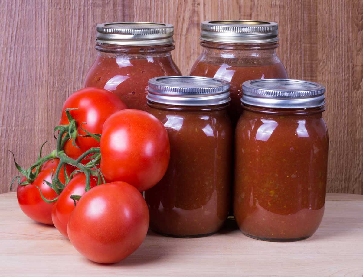 A close up of a set of glass jars with metal tops containing red tomato sauce five bright red tomatoes still attached to the vine.