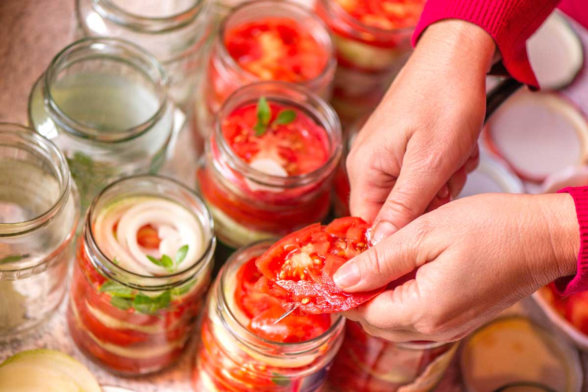 A close up of hands from the right of the frame chopping up tomatoes and placing them in jars for pickling.