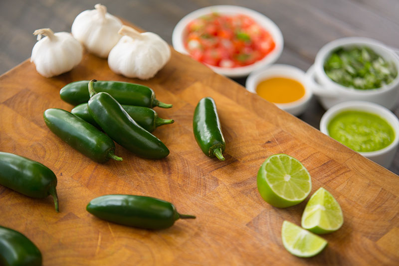 A close up of a wooden chopping board with sliced lime, 'Jalapeno' peppers, and garlic. In the background is condiments in white ceramic pots in soft focus.