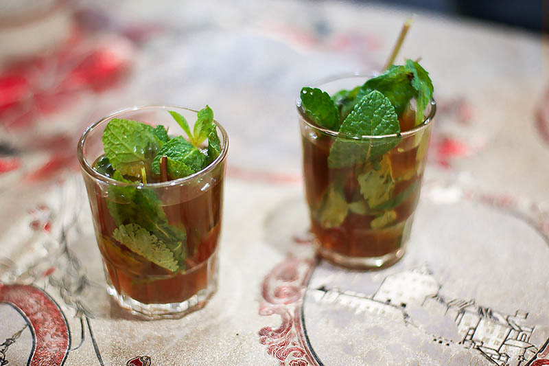 Two glasses containing cocktails garnished with mint set on a patterned table cloth on a soft focus background.