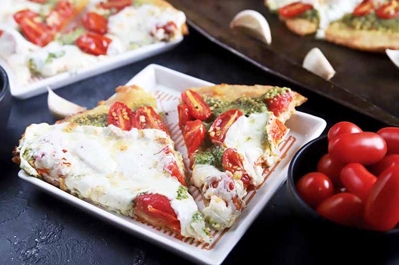 A close up of a white square dish with two slices of pizza with parsley pesto and mozzarella cheese. To the right of the frame is a small bowl of cherry tomatoes.