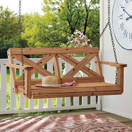 A close up of a wooden hanging bench seat on a wooden deck with a white wall to the right of the frame and a garden scene in the background.