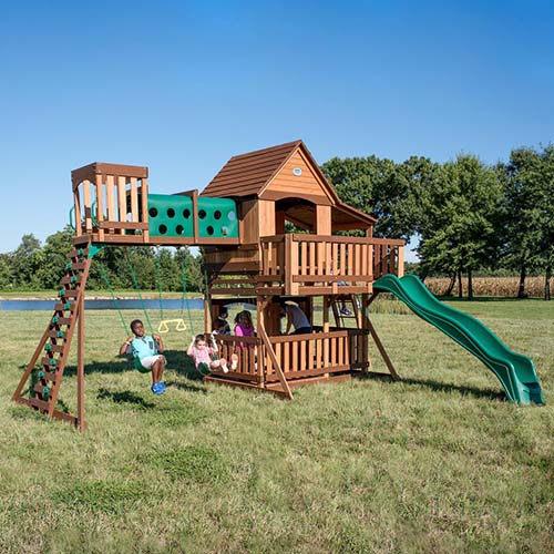A picture of the children's playset Woodridge II by Backyard Discovery, set up on a lawn with a lake and blue sky in the background.