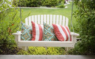 A close up of a white hanging bench overlooking a summer garden scene with lawns and shrubs in the background.