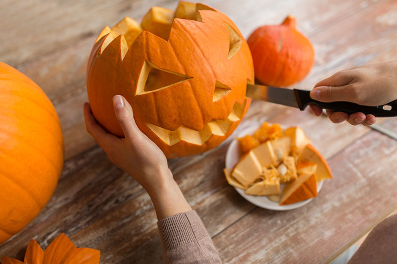 A close up of two hands from the right of the frame holding a large orange pumpkin, and using a knife to carve a face for Halloween.